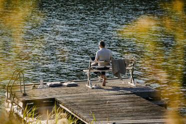 Rückansicht eines Mannes, der einen Laptop benutzt, während er auf einer Bank über einem Steg am See sitzt - MASF15723