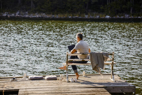 Älterer Mann, der einen Laptop benutzt, während er auf einer Bank über einem Holzsteg am See sitzt - MASF15727