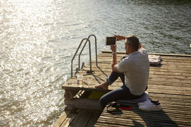 High angle view of mature male photographing on digital tablet while sitting on pier over lake - MASF15728