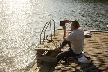 Reifer Mann fotografiert auf digitalem Tablet, während er auf einem Steg über dem See sitzt - MASF15728