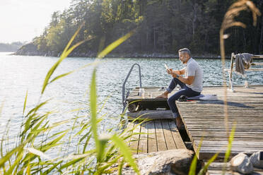 Full length of man using digital tablet while sitting on wooden pier over lake at forest - MASF15730