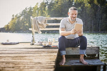 Smiling mature man using digital tablet with in-ear headphones while sitting on jetty over lake - MASF15734