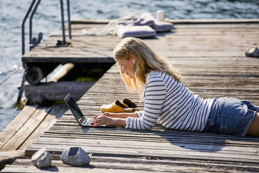 Mid adult female typing on laptop while lying over wooden pier during summer vacation - MASF15735