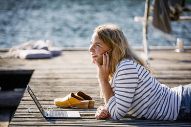 Smiling female with laptop talking on smart phone while lying over wooden pier - MASF15736