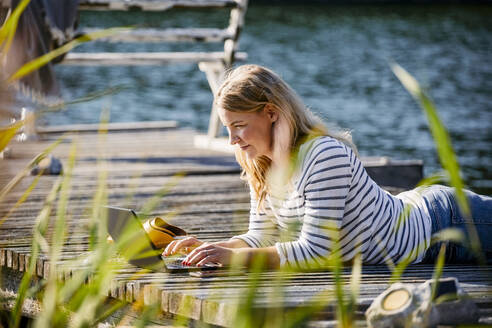 Frau auf der Vorderseite liegend bei der Arbeit über einem Laptop an der Seebrücke im Sommer - MASF15737