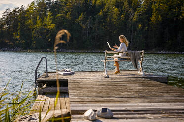 Mid adult woman doing video call on laptop while sitting on bench over wooden pier against lake in forest - MASF15738