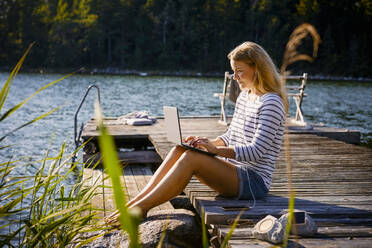 Frau mit Kopfhörern, die einen Laptop benutzt, während sie auf einem Steg am See sitzt - MASF15740