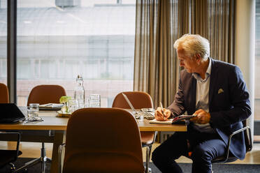 Älterer Jurist, der in sein Tagebuch schreibt, während er einen Laptop am Konferenztisch im Sitzungssaal benutzt - MASF15753