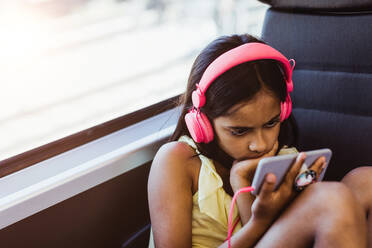 Girl wearing pink headphones watching movie on mobile phone while sitting by window in train - MASF15776