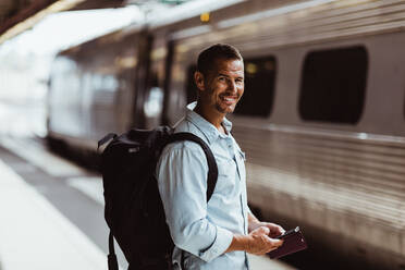 Porträt eines lächelnden Touristen mit Rucksack und Smartphone auf dem Bahnsteig eines Bahnhofs - MASF15788