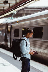 Mittlerer erwachsener Mann mit Rucksack, der ein Mobiltelefon benutzt, während er auf dem Bahnsteig neben dem Zug steht - MASF15791