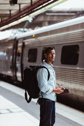 Thoughtful man with backpack holding smart phone while looking at train on platform - MASF15792