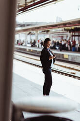 Businesswoman using mobile phone while standing at railroad station platform - MASF15828