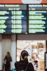 Rückansicht einer Geschäftsfrau, die ihr Smartphone in der Hand hält, während sie auf die Ankunft-Abfahrtstafel am Bahnhof schaut - MASF15830
