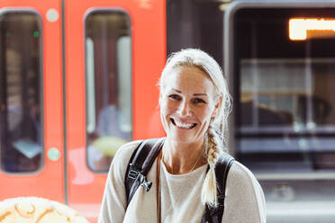 Porträt einer lächelnden reifen Frau mit Rucksack auf einem Bahnhof - MASF15835
