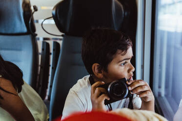 Neugieriger Junge mit Kamera, der im Zug sitzend das Fenster betrachtet - MASF15842
