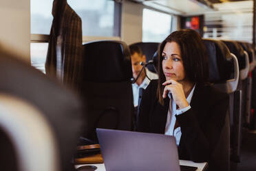 Thoughtful business commuter using laptop while looking away in train - MASF15856