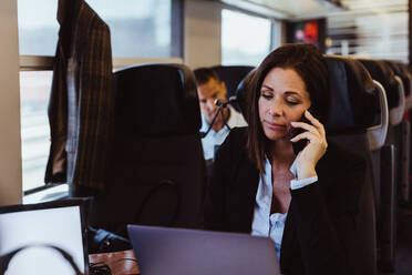 Businesswoman talking on smart phone and using laptop while commuting in train - MASF15857