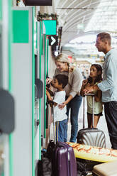 Mutter, die ihrem Sohn beim Kauf einer Fahrkarte hilft, während die Familie mit Gepäck auf dem Bahnsteig steht - MASF15866