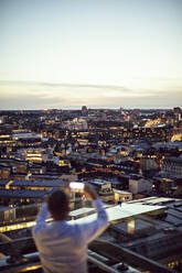 High angle view of businessman photographing illuminated city on mobile phone from office terrace during sunset - MASF15876