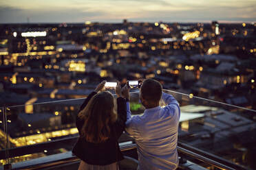 Rear view of male and female professionals photographing city on mobile phone from terrace during sunset - MASF15877