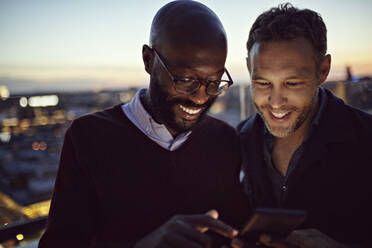 Smiling male professionals looking at mobile phone while standing on terrace during sunset - MASF15882