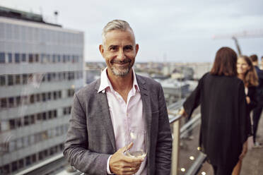 Porträt eines lächelnden Geschäftsmannes, der ein Weinglas hält, mit Mitarbeitern im Hintergrund auf einer Terrasse - MASF15895