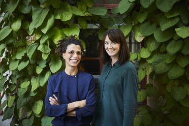 Portrait of smiling female architects standing against creeper plants in backyard - MASF15949