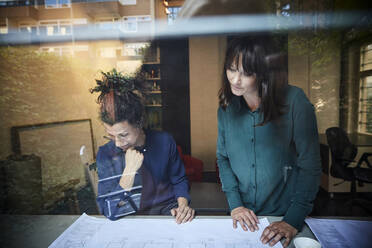 Female architects discussing over blueprint at table seen through glass in office - MASF15965