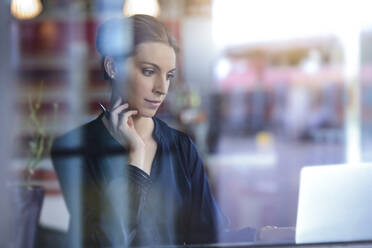 Businesswoman using laptop behind windowpane in a cafe - BMOF00124