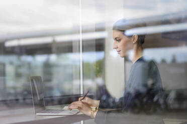 Businesswoman using laptop behind windowpane in a cafe - BMOF00126