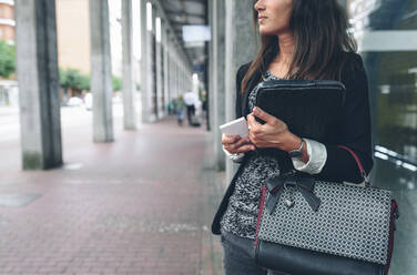 Midsection of woman holding mobile phone with purse standing by window - CAVF70991