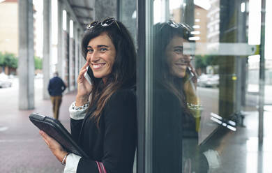 Porträt einer glücklichen Frau, die am Fenster stehend mit ihrem Handy telefoniert - CAVF70993