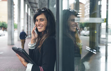 Portrait of happy woman talking on mobile phone while standing by window - CAVF70993