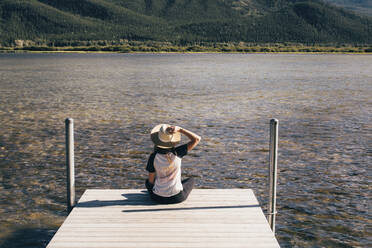 Rear view of woman sitting on jetty by lake - CAVF71007