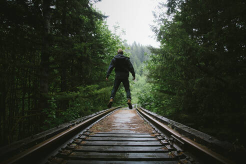 Rückansicht eines Mannes, der auf Eisenbahnschienen inmitten von Bäumen springt - CAVF71018
