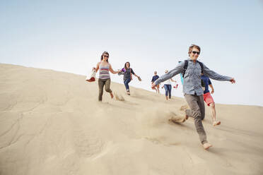 Cheerful friends running down sand dune against clear sky - CAVF71056