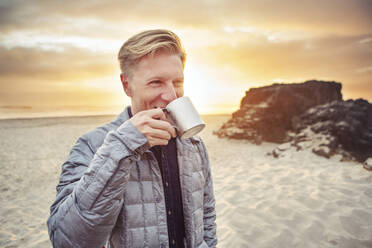 Glücklicher Mann trinkt Alkohol aus einem Becher, während er bei Sonnenuntergang am Strand steht - CAVF71078