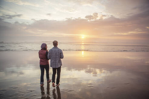 Rear view of couple walking at beach against sky during sunset - CAVF71083
