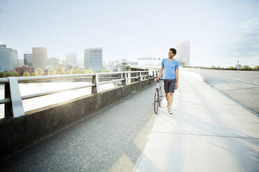 Male athlete walking with bicycle on footpath in city - CAVF71143