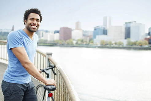 Porträt eines männlichen Sportlers auf einer Brücke mit Fahrrad am Fluss - CAVF71147