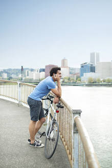 Männlicher Sportler, der ein Mobiltelefon benutzt, während er mit dem Fahrrad auf einer Brücke am Fluss steht - CAVF71148