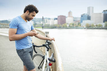 Sportler, der sein Smartphone benutzt, während er mit dem Fahrrad auf einer Brücke am Fluss steht - CAVF71149