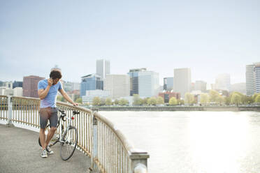 Young athlete using mobile phone while standing with bicycle on bridge by river - CAVF71150