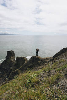 Rückansicht eines Mannes, der auf einem Berg steht und auf das Meer blickt, vor einem bewölkten Himmel - CAVF71173
