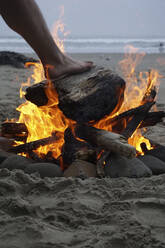 Abgeschnittenes Bein eines Mannes auf brennendem Brennholz am Strand - CAVF71179