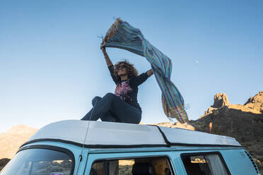 Low angle view of happy woman with arms raised holding scarf while sitting on travel trailer against clear sky - CAVF71249