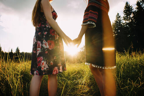Midsection of lesbian couple holding hands while standing on grassy field during sunset - CAVF71255