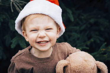 Portrait of happy boy with teddy bear wearing Santa hat in backyard - CAVF71277