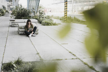 Young smiling woman sitting on the ground and using tablet - KNSF06970